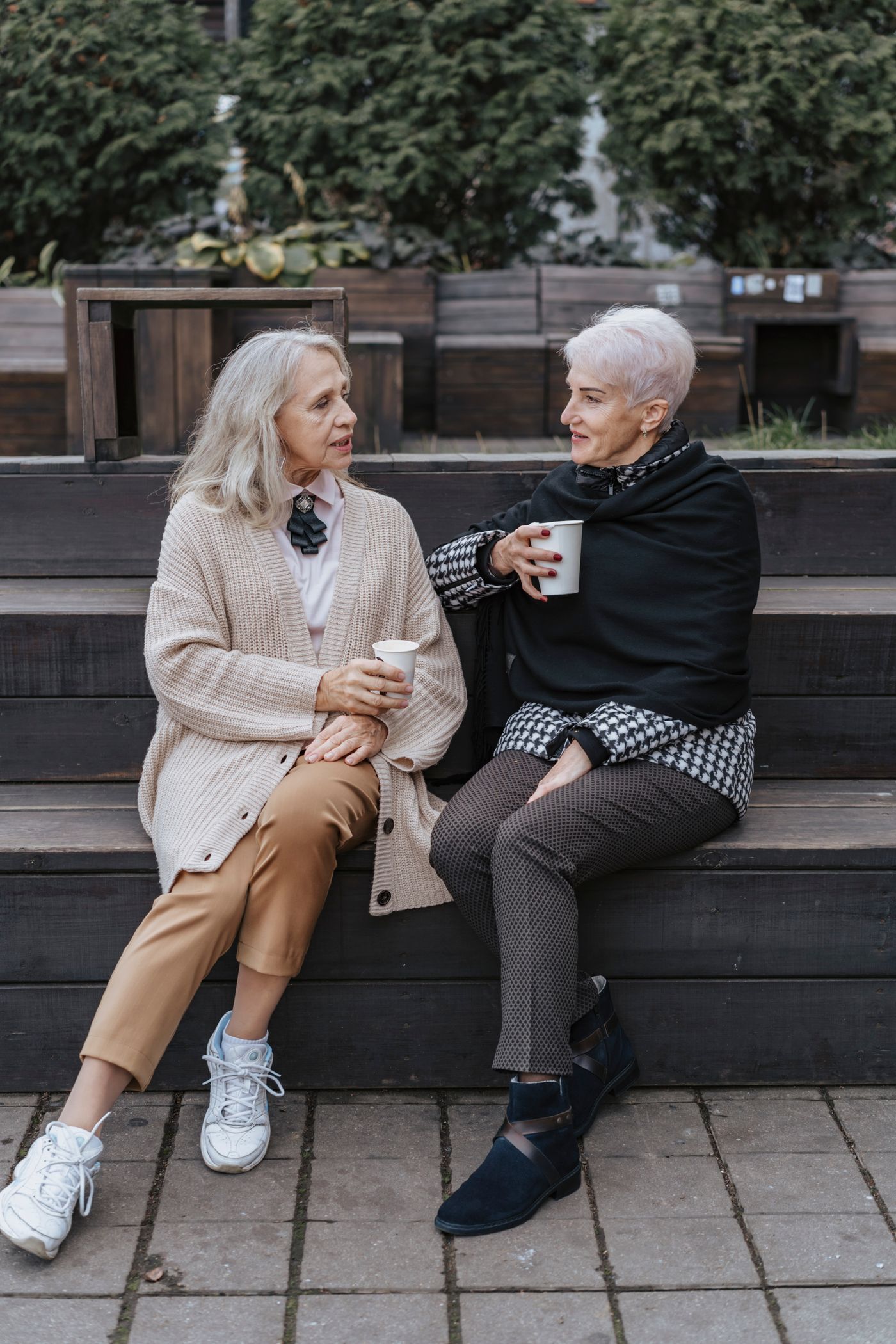 Two older women sitting on a bench with warm drinks